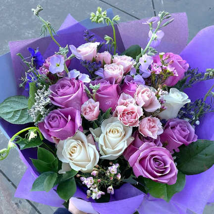 Hand-tied bouquet of lavender, pink, and white roses with accent flowers wrapped in purple paper.