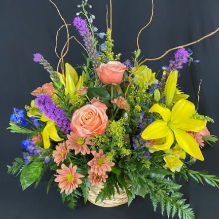 Basket arrangement with peach roses, yellow lilies, purple spikes, and blue flowers.