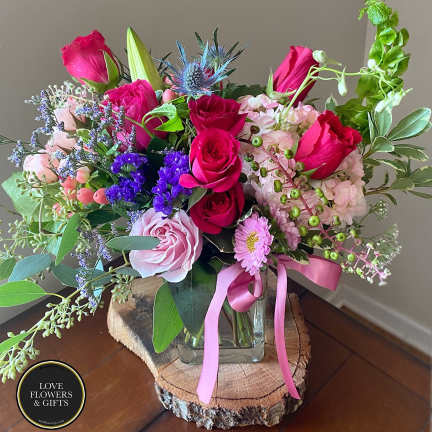 Pink and purple mixed flower arrangement with roses and hydrangea in a glass vase on a wood slice stand.