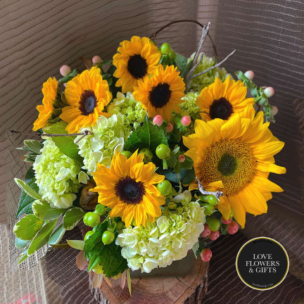 Arrangement of yellow sunflowers and pale green hydrangeas in a low vase on a wood slice