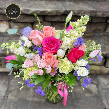 Mixed bouquet of pink, white, and purple flowers arranged in a clear glass vase with a pink ribbon.