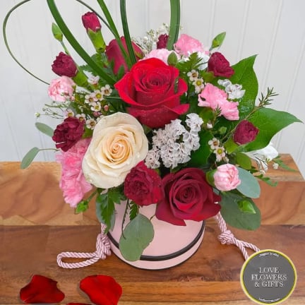 Pink hatbox arrangement with red and cream roses, pink carnations, and decorative grass loops on a wooden surface.