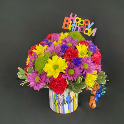 Colorful mixed flower arrangement in a striped birthday container with a Happy Birthday topper
