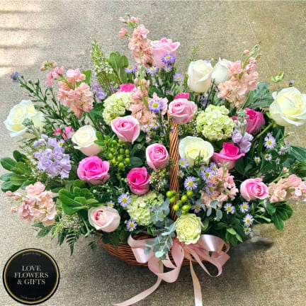 Basket arrangement of pink and white roses with pastel filler flowers and a pink ribbon