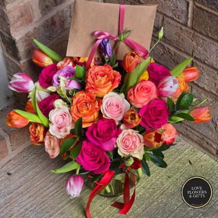 Bright bouquet of pink roses and orange tulips in a glass vase with a kraft card tied with pink ribbon.