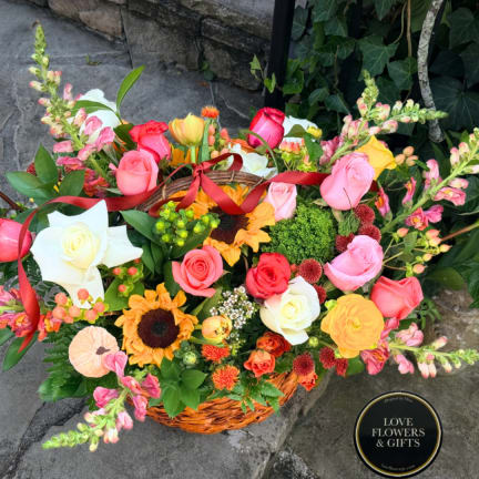 Basket arrangement of roses, sunflowers, and snapdragons with a red ribbon