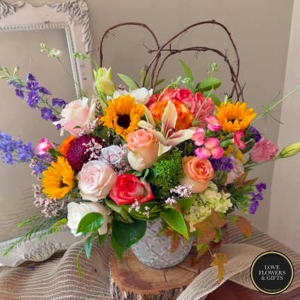 Colorful arrangement of sunflowers, roses, and lilies in a white ceramic pot with twig accents.