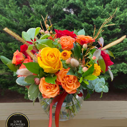 Round glass vase of yellow, orange, and red roses with small yellow mums and berries, tied with an orange ribbon