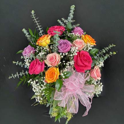 Mixed arrangement of pink, peach, lavender, and yellow roses with white filler flowers and a pink ribbon bow