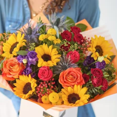Mixed bouquet with sunflowers, roses, and purple blooms wrapped in orange paper