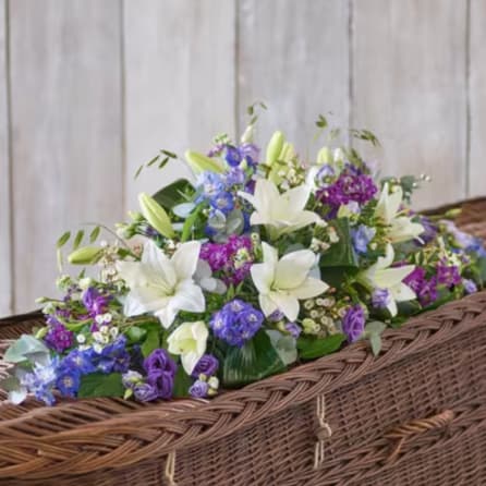 White lilies and purple-blue flowers arranged in a wicker casket spray