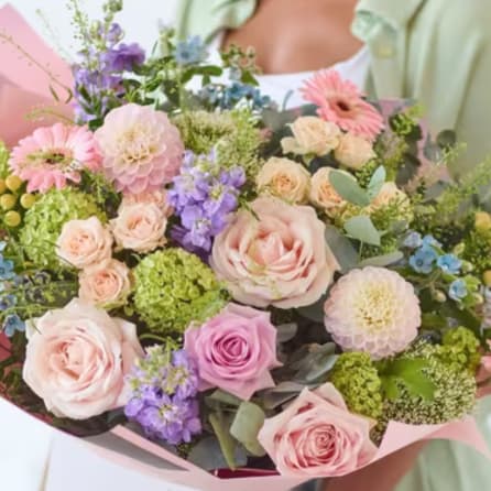Pastel bouquet of roses, dahlias, and purple blooms wrapped in pink paper