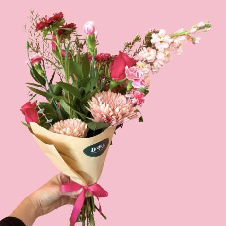 Handheld bouquet of pink and red flowers wrapped in tan paper with a pink ribbon