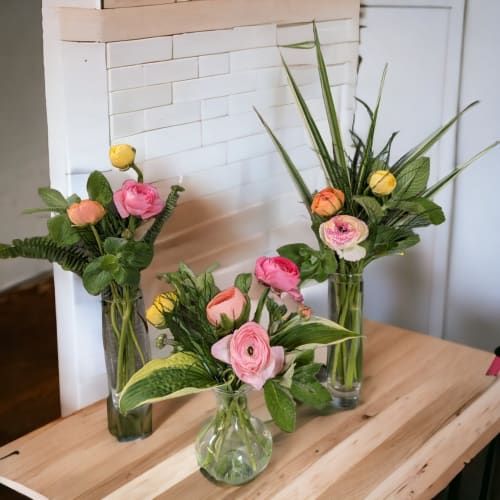 Three small arrangements of pink and yellow ranunculus in clear glass vases on a wooden table