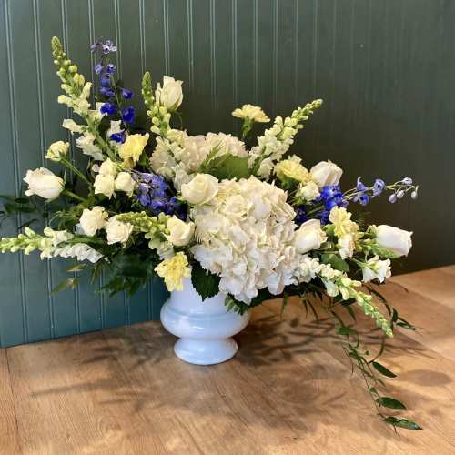 Wide white pedestal vase with white hydrangeas, roses, yellow blooms, and blue delphinium in an asymmetrical design
