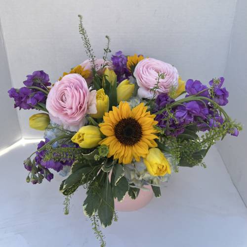 Bouquet with pink ranunculus, yellow tulips, purple flowers, and a sunflower in a pink container