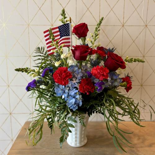 Patriotic arrangement with red roses, red carnations, blue hydrangeas and a small American flag in a white vase