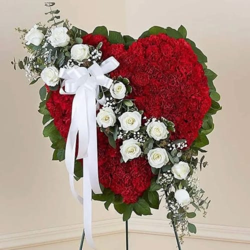 Standing heart-shaped spray of red carnations with white roses and a white ribbon on an easel.