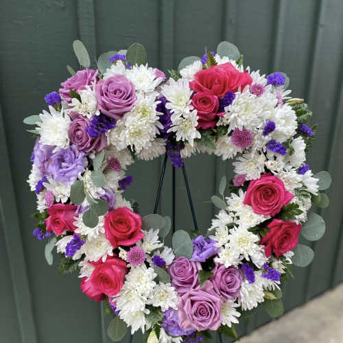 Heart-shaped wreath of pink and lavender roses with white mums and purple accents on a standing easel.