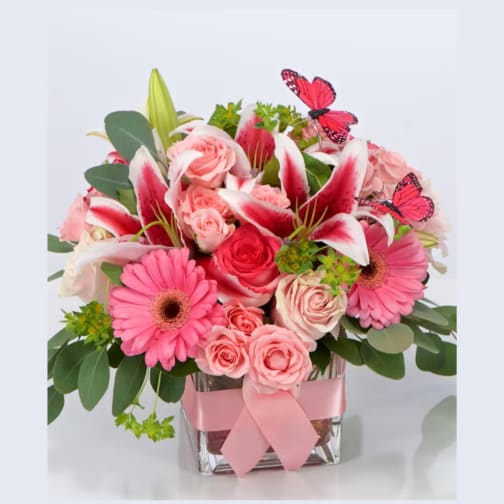 Pink lilies, roses, and gerbera daisies in a glass cube vase with pink ribbon and butterfly accents