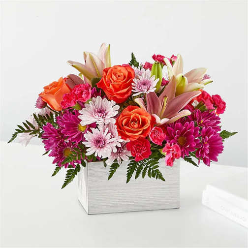 Low rectangular arrangement of orange roses, pink lilies, and mixed pink and white mums in a white box vase