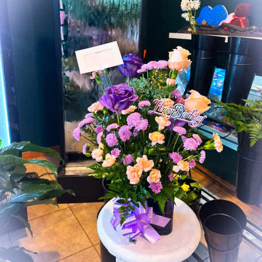 Purple and peach flower arrangement with a Happy Birthday sign in a black container