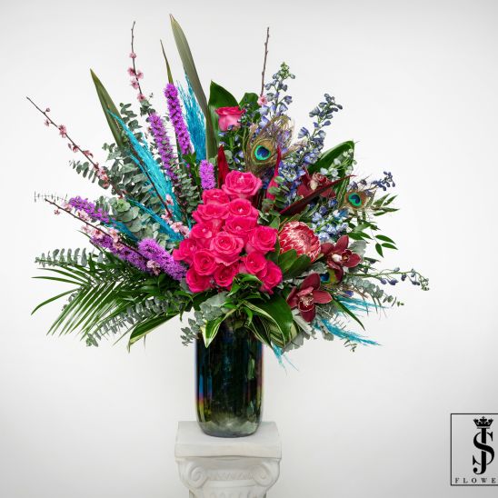 Tall arrangement of hot pink roses, orchids, protea, and peacock feathers in a dark glass vase