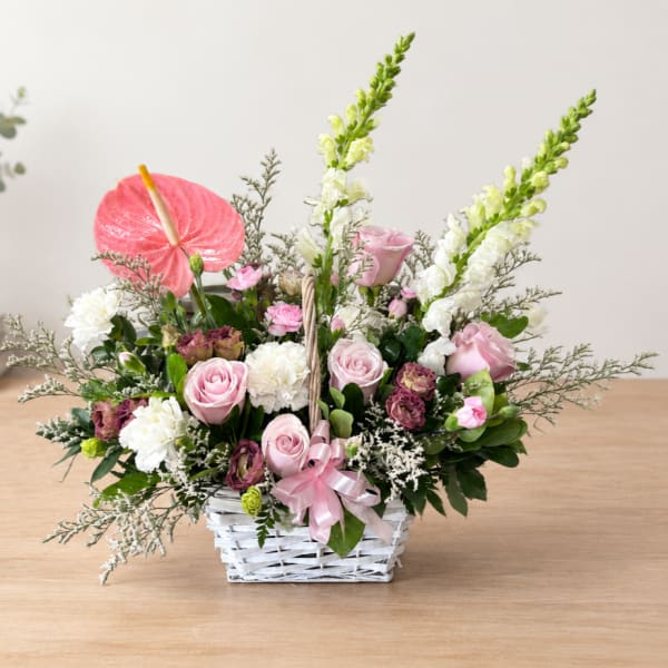 Pink roses and white flowers arranged in a white basket with a ribbon.