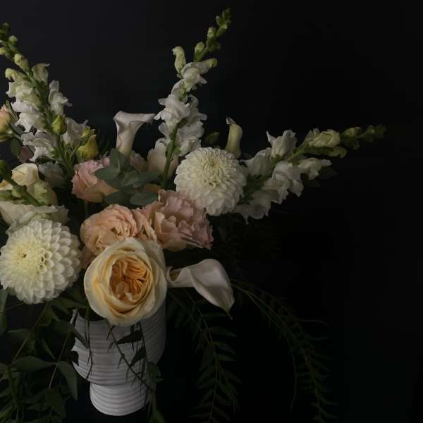 Soft peach and white flowers arranged in a white vase against a dark background.