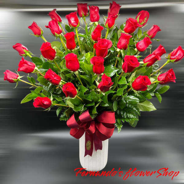 Tall white vase filled with many red roses and foliage, finished with a large red satin bow