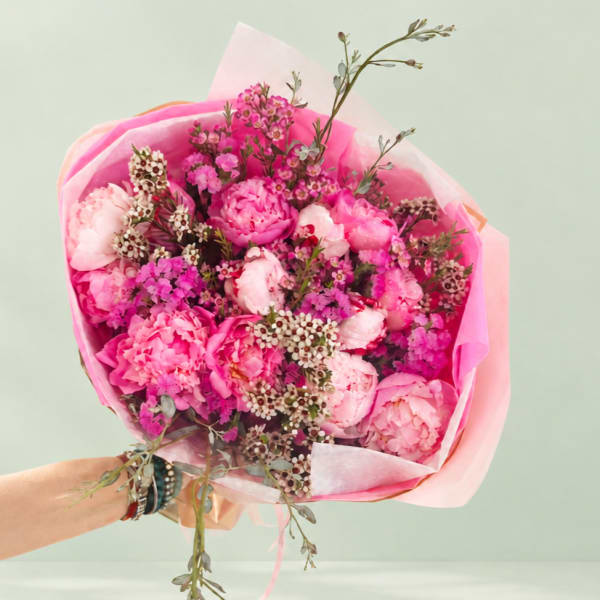 Handheld bouquet of pink peonies and small filler flowers wrapped in pink paper
