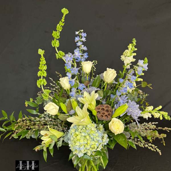 Tall floral arrangement in a glass vase with white roses, blue flowers, and lilies