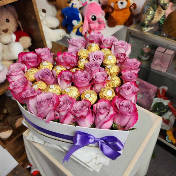 Pink roses arranged with gold-wrapped chocolates in a white box with a purple ribbon