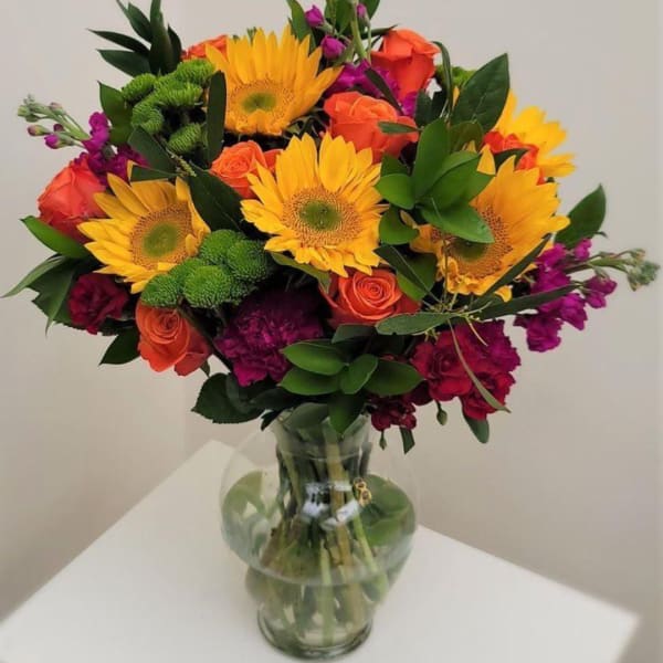 Bouquet of sunflowers, orange roses, and purple flowers in a clear glass vase