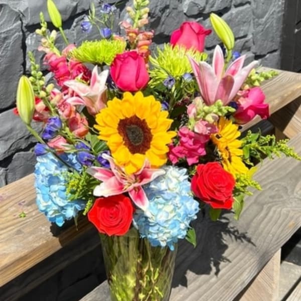 Colorful mixed bouquet in a clear glass vase with sunflowers, roses, lilies, and hydrangeas.