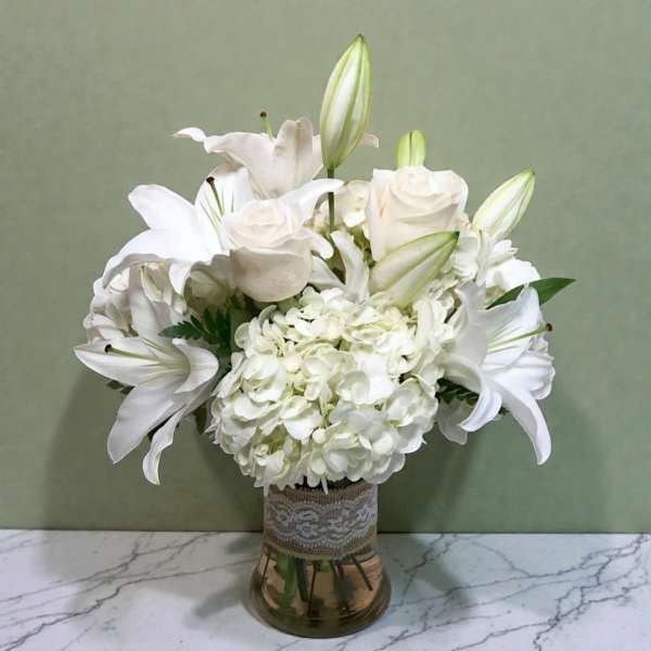 White lilies and hydrangeas arranged in a glass vase with lace trim
