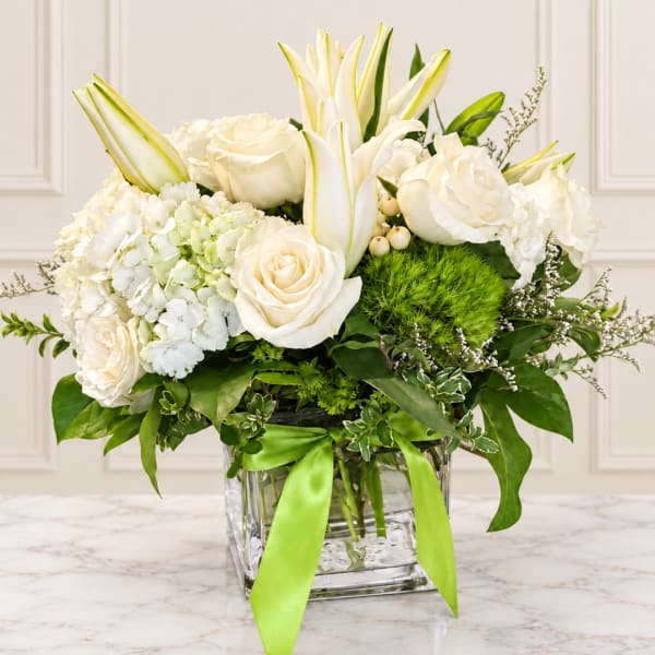 White roses and lilies arranged in a clear glass vase with green ribbon
