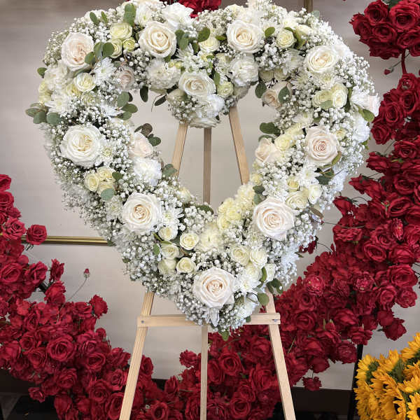 Heart-shaped white rose wreath on a wooden easel, surrounded by red roses