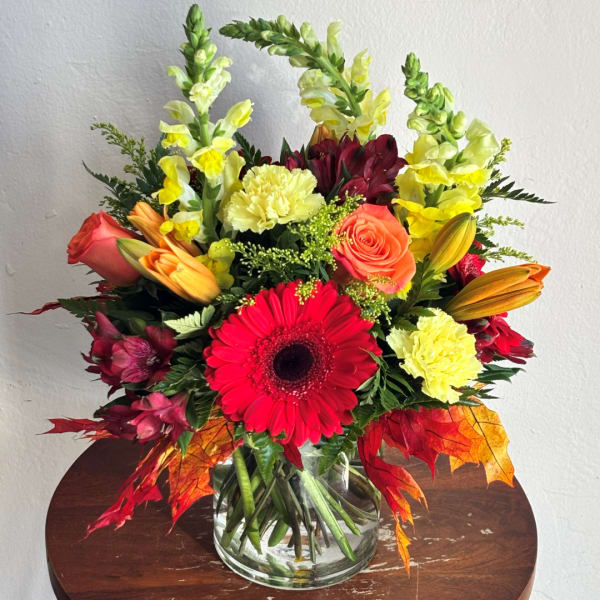 Mixed bouquet in a clear glass vase with red gerbera, roses, lilies, and yellow snapdragons.