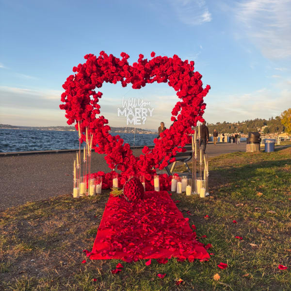 Heart-shaped rose arch with a red petal aisle and proposal sign by the water