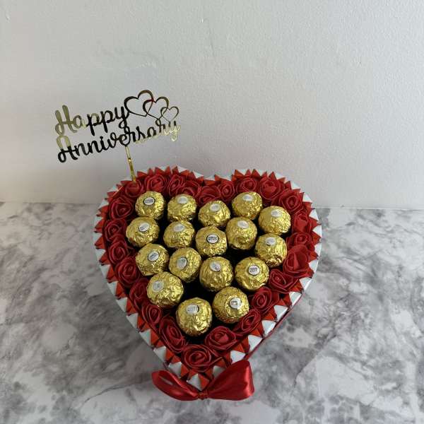 Heart-shaped box of red roses and gold-wrapped chocolates with a Happy Anniversary topper