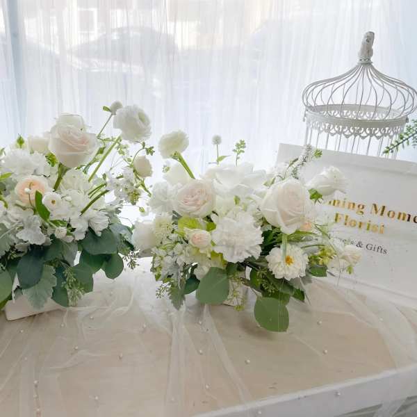 Two white floral arrangements on a table with a decorative birdcage