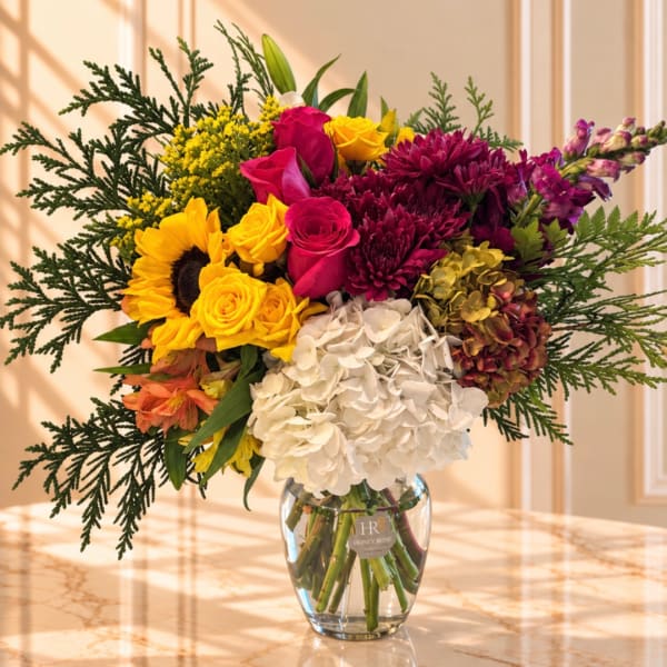 Colorful mixed bouquet in a clear glass vase with roses, sunflowers, hydrangea, and orchids