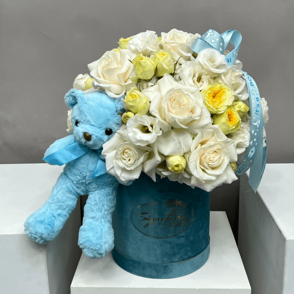 White and pale yellow roses in a blue hatbox with a blue teddy bear