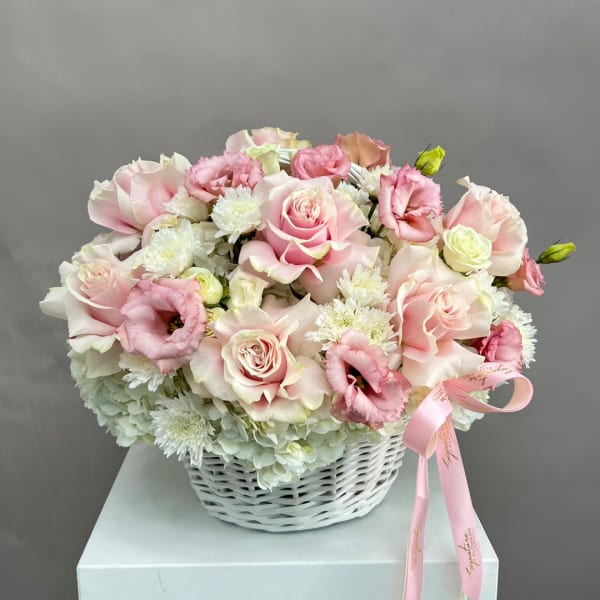Pink roses and white blooms arranged in a white basket with a pink ribbon