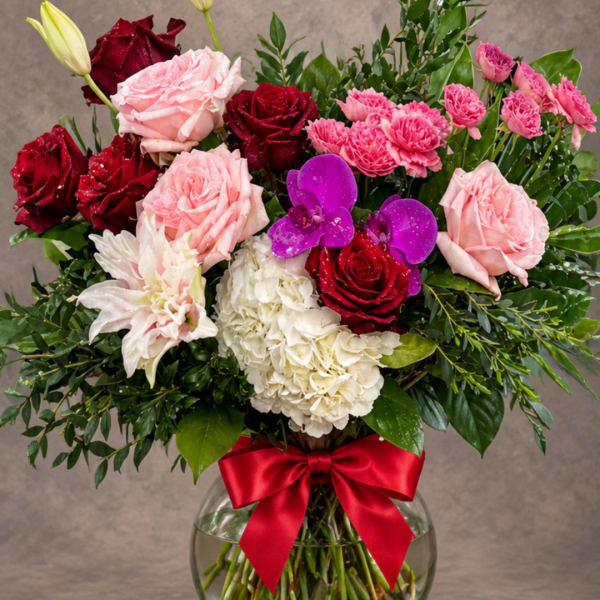 Mixed bouquet of roses, hydrangea, and orchids in a glass vase with a red bow
