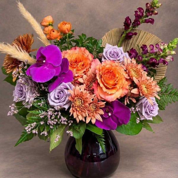 Mixed bouquet of orange, lavender, and purple flowers in a dark glass vase