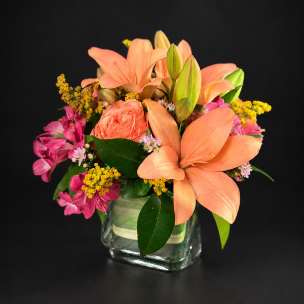 Peach lilies and pink flowers arranged in a square glass vase