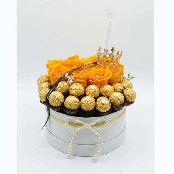 Orange roses in a round gift box with gold-wrapped chocolates and a crown topper