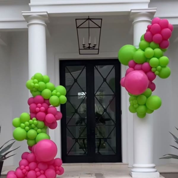 Pink and green balloon clusters frame a black front door on a white porch.